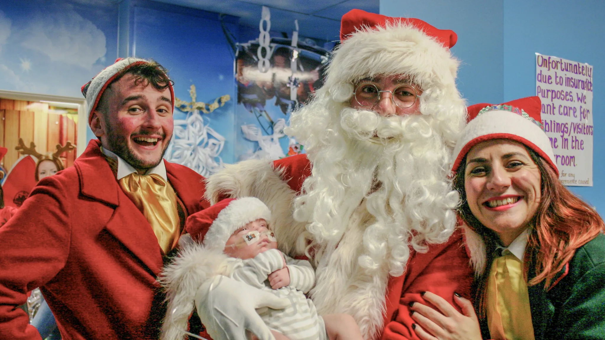 A mum and two children interact with a large costumed character dressed as Santa Claus in bright red with a white beard and hat. The setting appears to be indoors, decorated for Christmas with a Christmas tree and wrapped presents visible in the background.