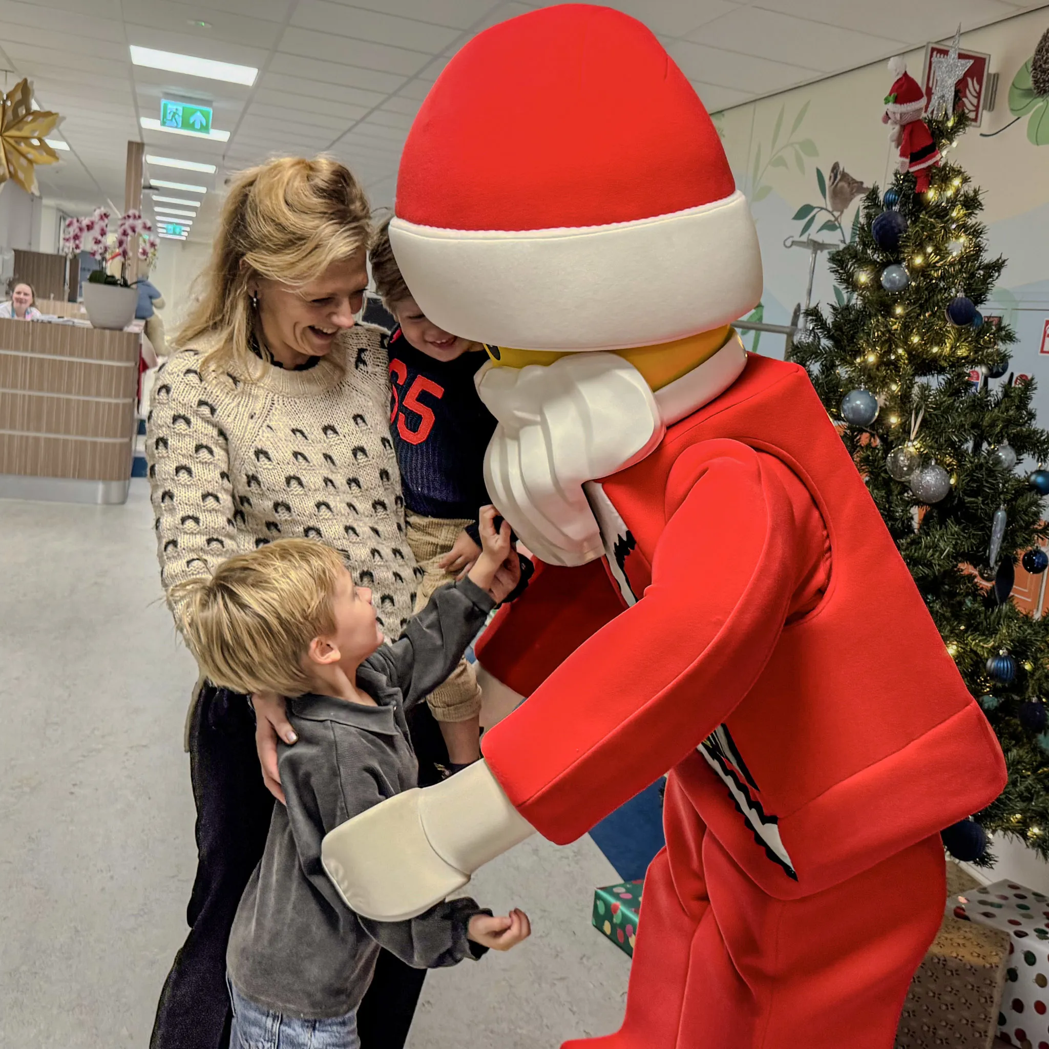 A person and two children interact with a large costumed character dressed as Santa Claus in bright red with a white beard and hat. The setting appears to be indoors, decorated for Christmas with a Christmas tree and wrapped presents visible in the background.