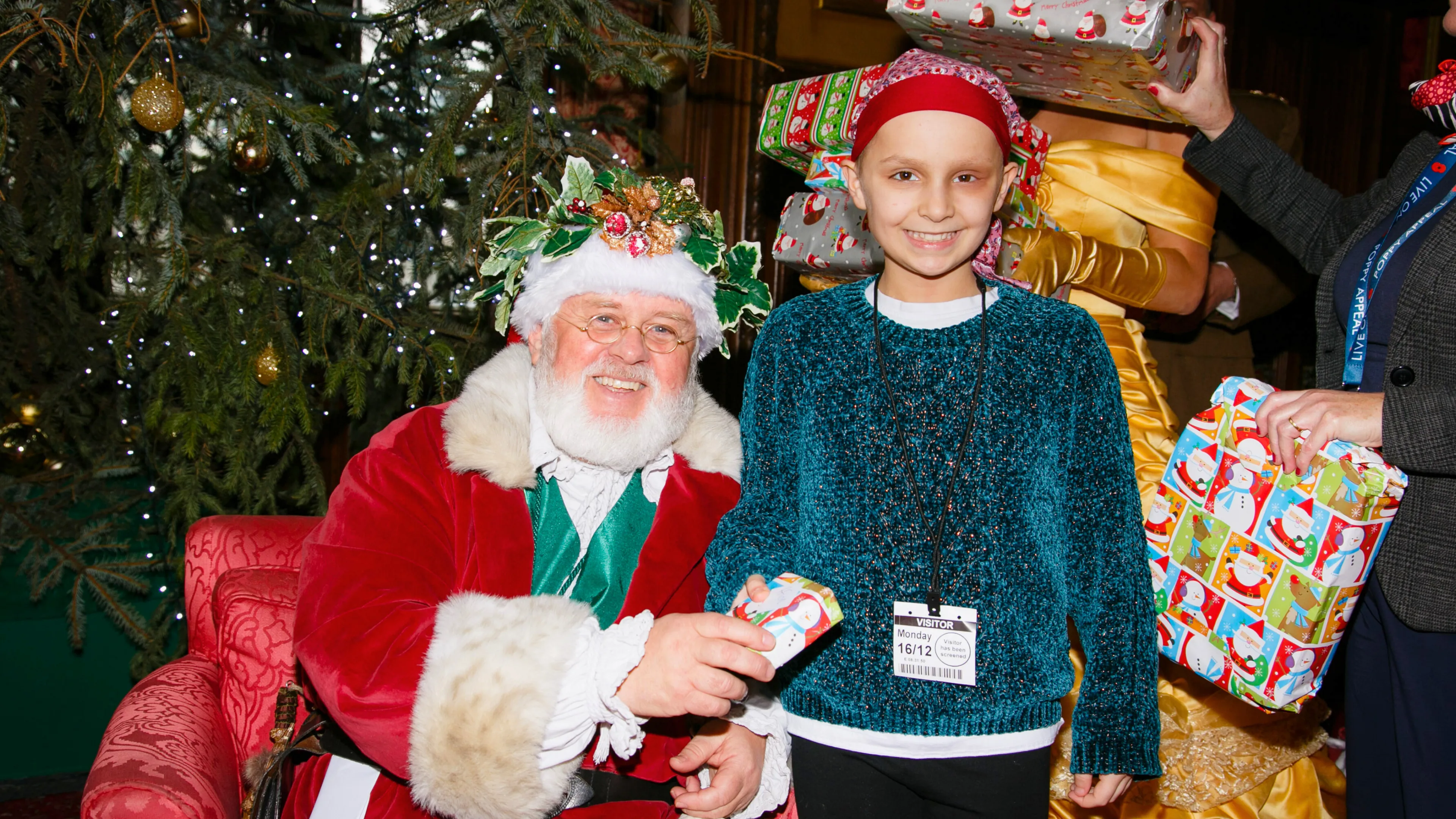 A smiling young child wearing a red headscarf and teal sweater stands beside Santa Claus, who is seated and handing the child a small wrapped gift. Behind them is a large decorated Christmas tree and people holding festive presents.