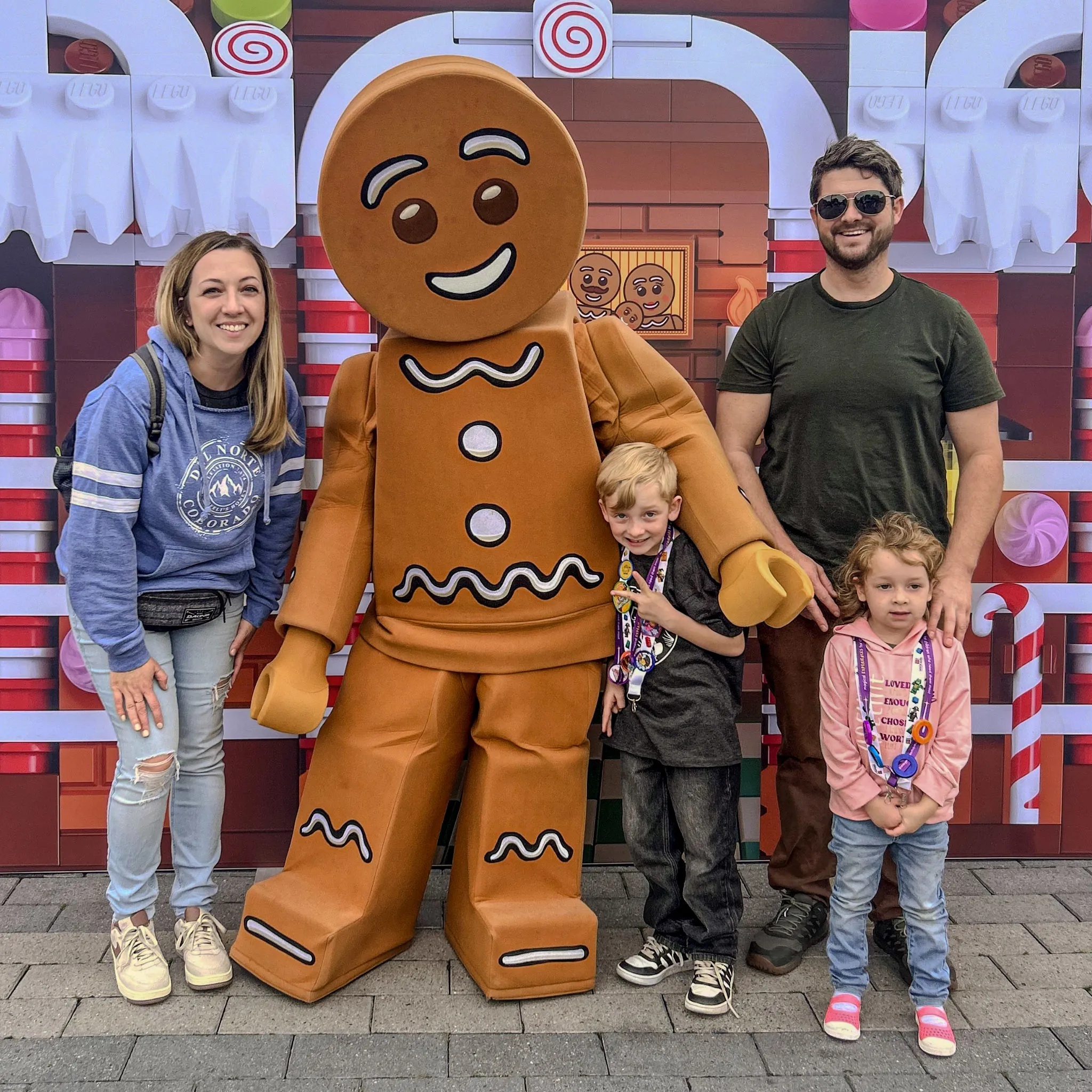 A family of four people pose with a large gingerbread man character in front of a festive backdrop featuring candy-themed decorations. The gingerbread figure has a smiling face and white icing details on its costume.